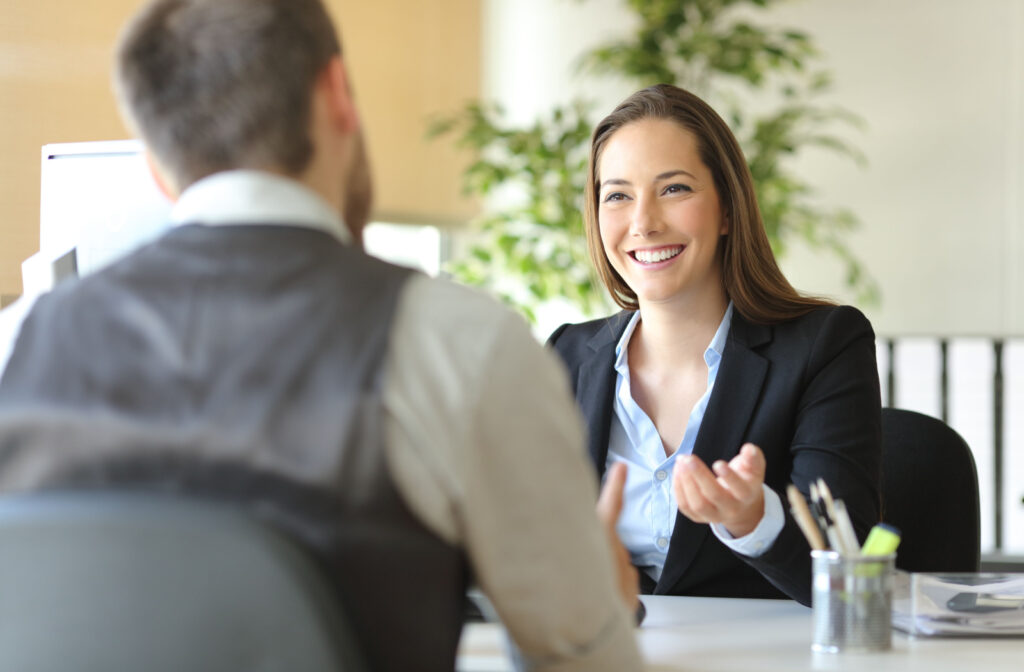 Two working professionals sitting at a table and talking. A woman is smiling and gesturing with her hands as a man sits facing her, out of focus.