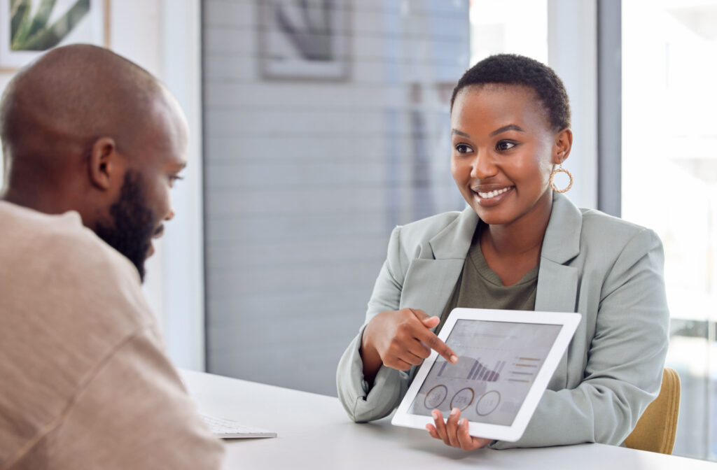 Two working professionals sitting at a table and talking. A woman is smiling and gesturing with her hands as a man sits facing her, out of focus.