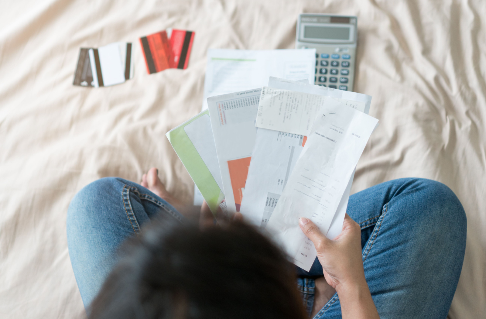 Close-up of a person's hands typing numbers into a calculator while holding a piece of paper. There is a stack of bills and credit cards spread out on the table.
