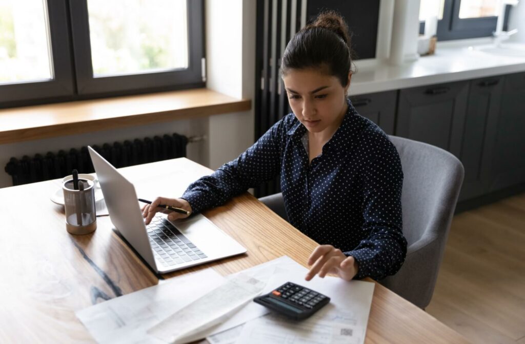 A person sitting at a desk and using their laptop, paper and pen, and a calculator to create a budget and plan for a short-term loan.