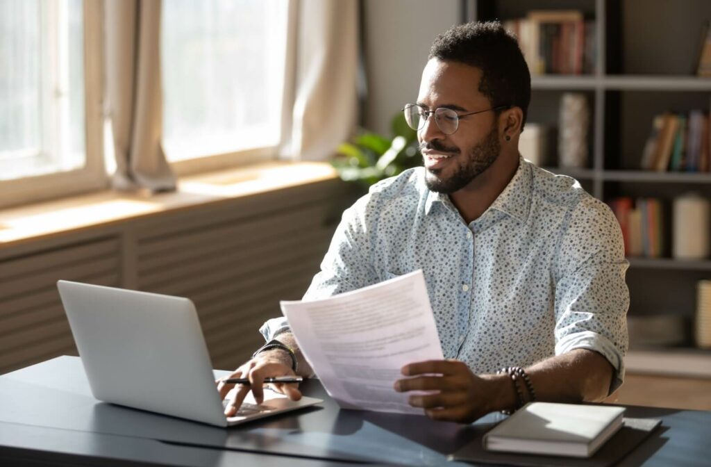 A person smiling as they use their laptop to fill out an online loan application and consult a piece of paper with costs and loans terms on it.